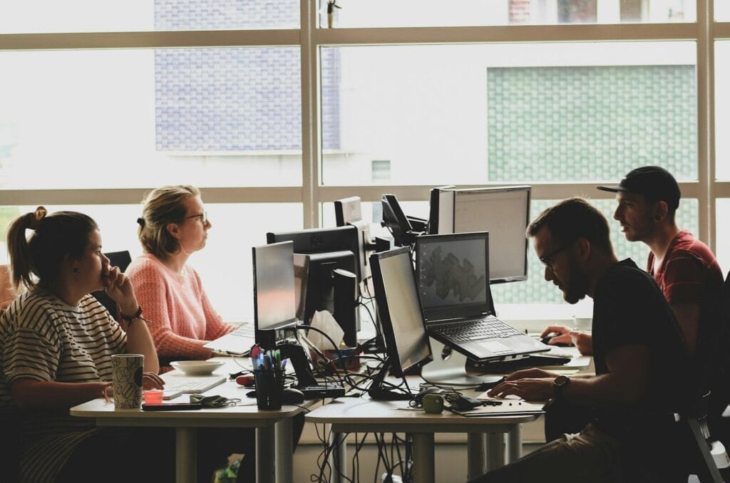 Four people working at computers in a sunlit office workspace