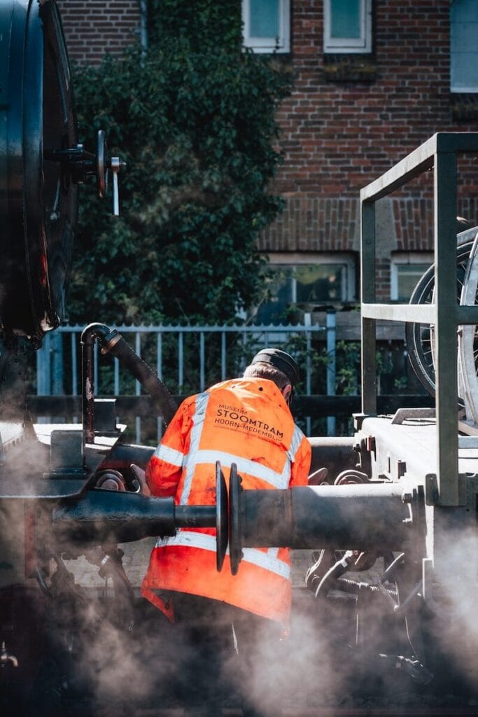 A worker repairs machinery by a brick building.